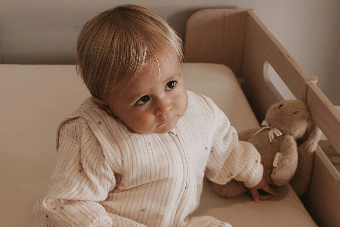 Bimbo nel lettino in legno con sacco nanna a righe e coniglietto di peluche, cameretta in stile nordico