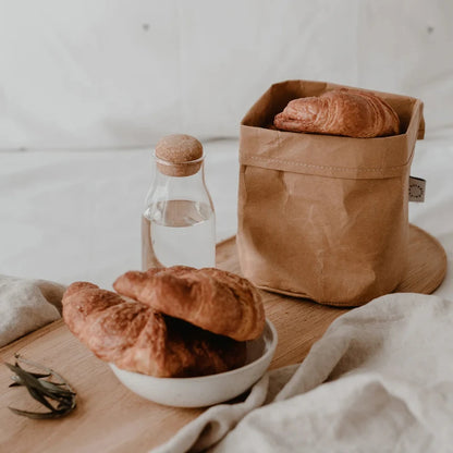 set di borse di carta lavabile marrone con croassaint e acqua su tagliere di legno e tovaglia di lino